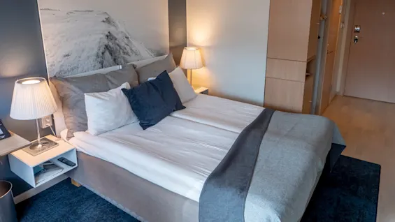 En prydligt bäddad hotellsäng med vita och grå sängkläder mot en mörk vägg med två lampor på sängbord. En naturskön bild hänger ovanför, i en mysig rumsmiljö.