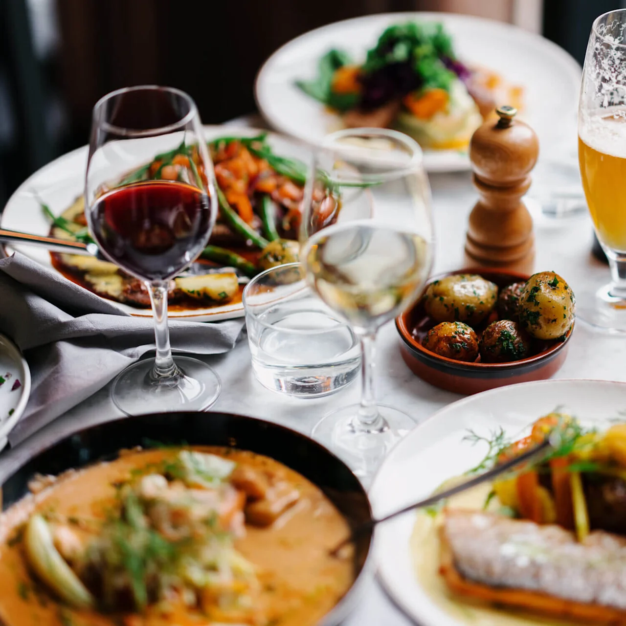 Plates of diverse dishes with wine and beer on a table, accented by a wooden pepper grinder, surrounded by a lively restaurant atmosphere.