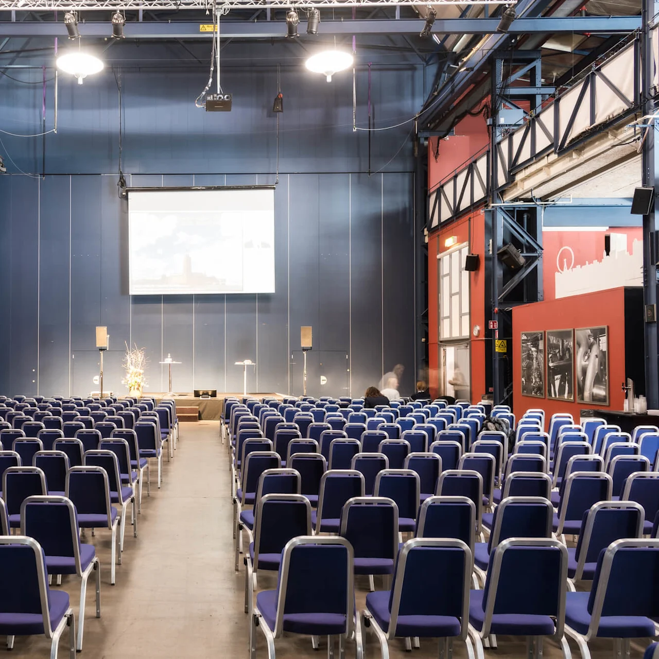 Rows of empty blue chairs face a screen in an industrial-style conference room with red and blue walls. Two spotlights illuminate the space, and a podium stands below the screen.