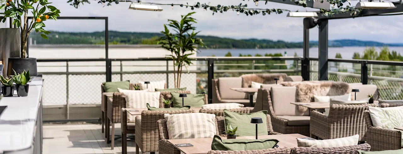 Kurvstoler med puter er arrangert under lyslenker på en takterrasse. Planter og utsikt mot et fjernt landskap kompletterer omgivelsene.