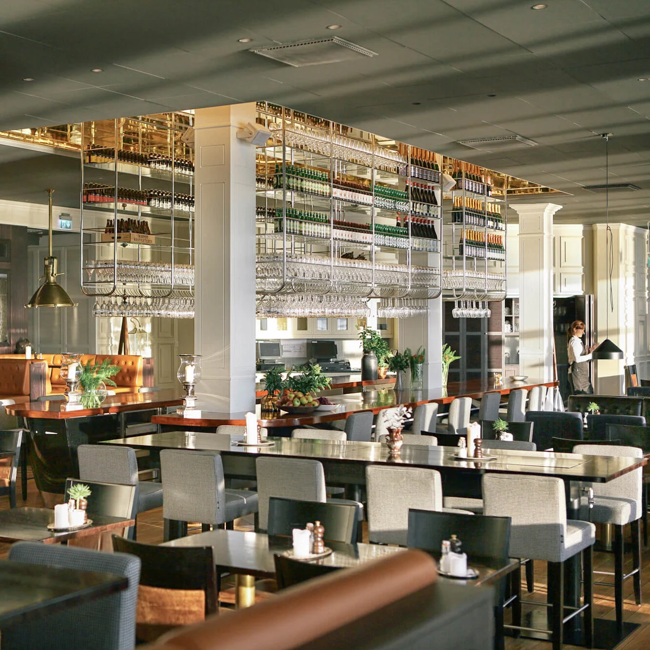 Restaurant interior with neatly arranged tables and chairs, hanging glasses above the bar, and a warmly lit ambiance. A person stands near the entrance. Lush greenery decorates the space.