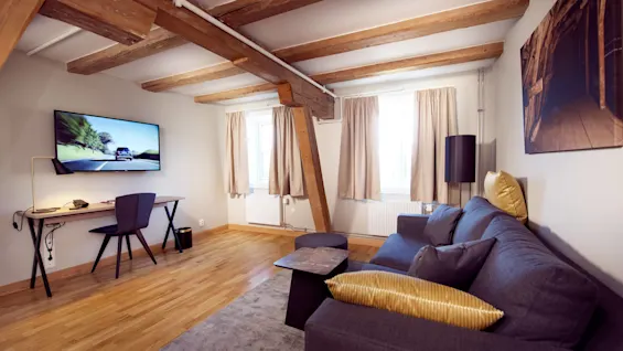 Et koselig rom med synlige trebjelker, en sofa, skrivebord med stol, og en flatskjerm-TV på veggen.