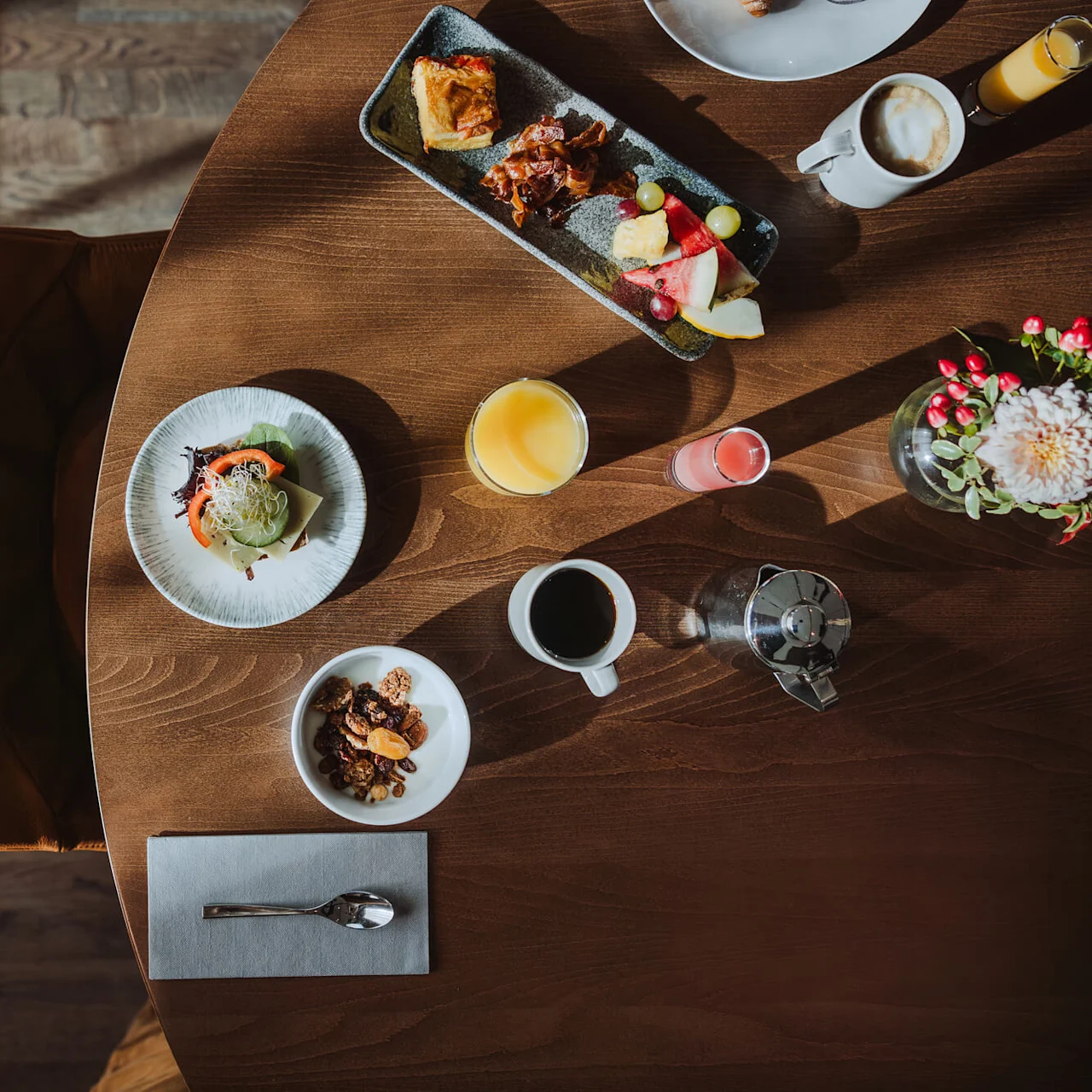 Ett runt träbord med en varierad frukost, inklusive en smörgås, juice, kaffe och frukt med blommor i närheten, i en solig matsal.