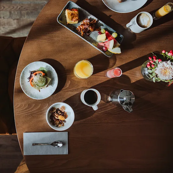 Et rundt trebord med en variert frokost, inkludert en sandwich, juice, kaffe og frukt med blomster i nærheten, i en solfylt spisestue.