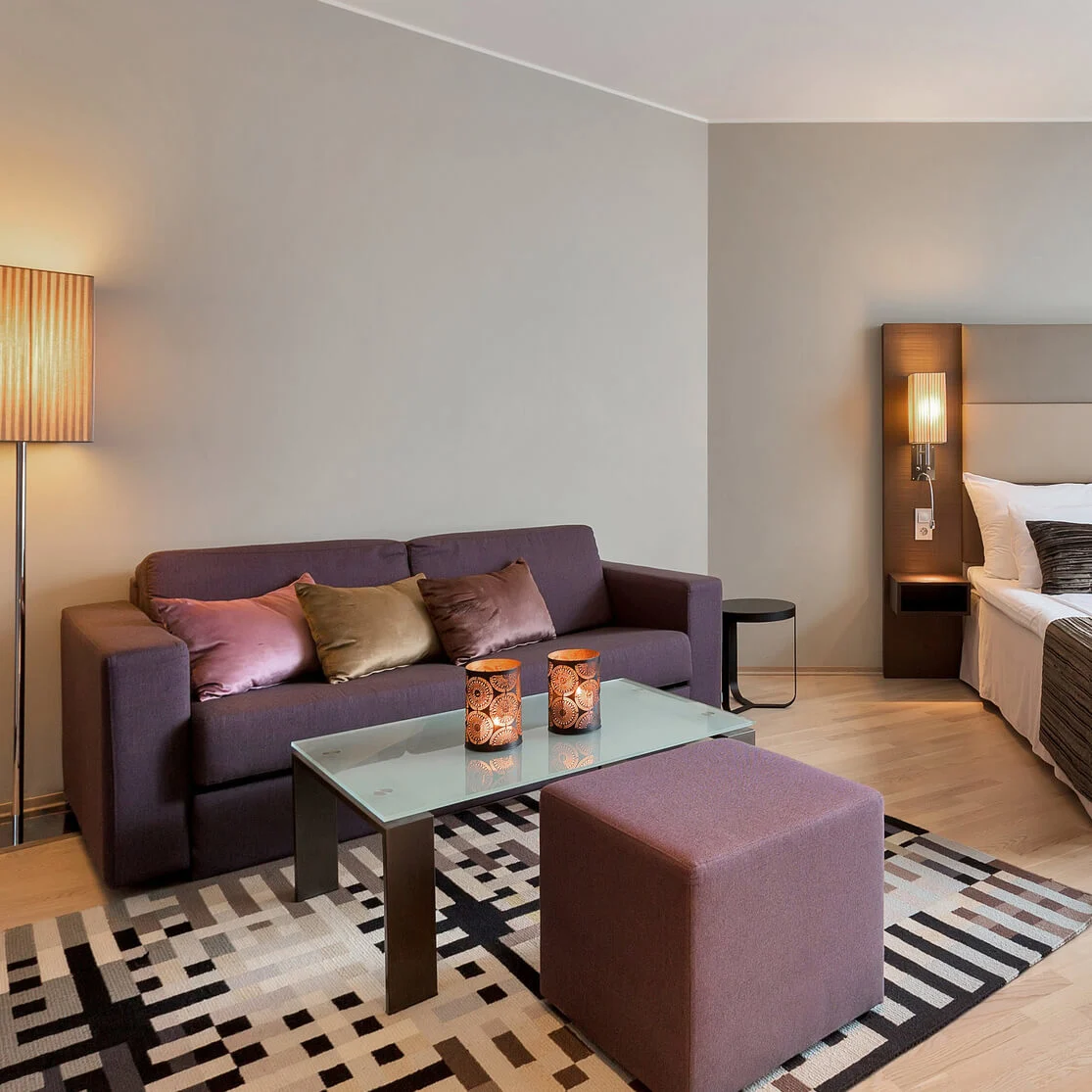 A hotel room with a large bed, patterned cushions, and a glass coffee table, featuring modern lighting and warm, neutral colors, creates a cozy and welcoming atmosphere.
