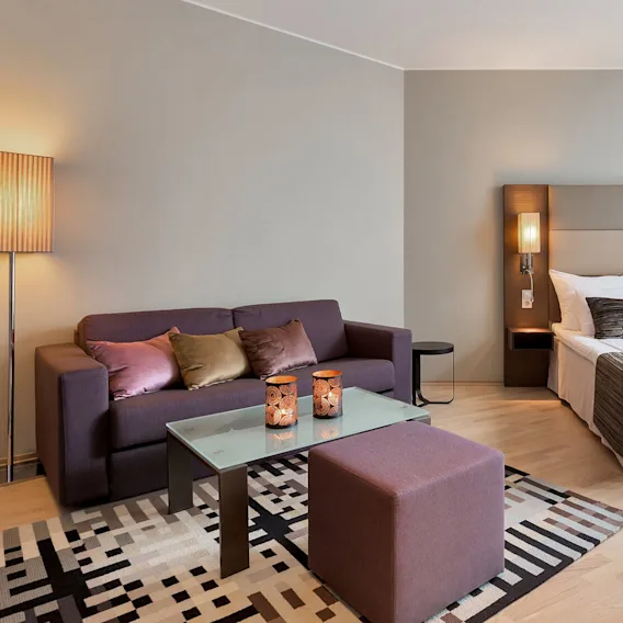 Et hotellrom med en stor seng, mønstrede puter og et glass sofabord, med moderne belysning og varme, nøytrale farger, skaper en koselig og innbydende atmosfære.