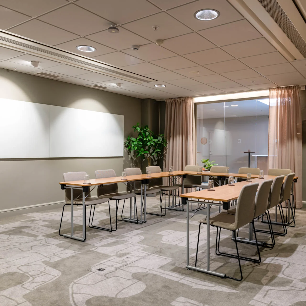 A conference room holds a U-shaped arrangement of tables with beige chairs and glass cups, set against a backdrop of a whiteboard and potted plants, under ceiling lights.