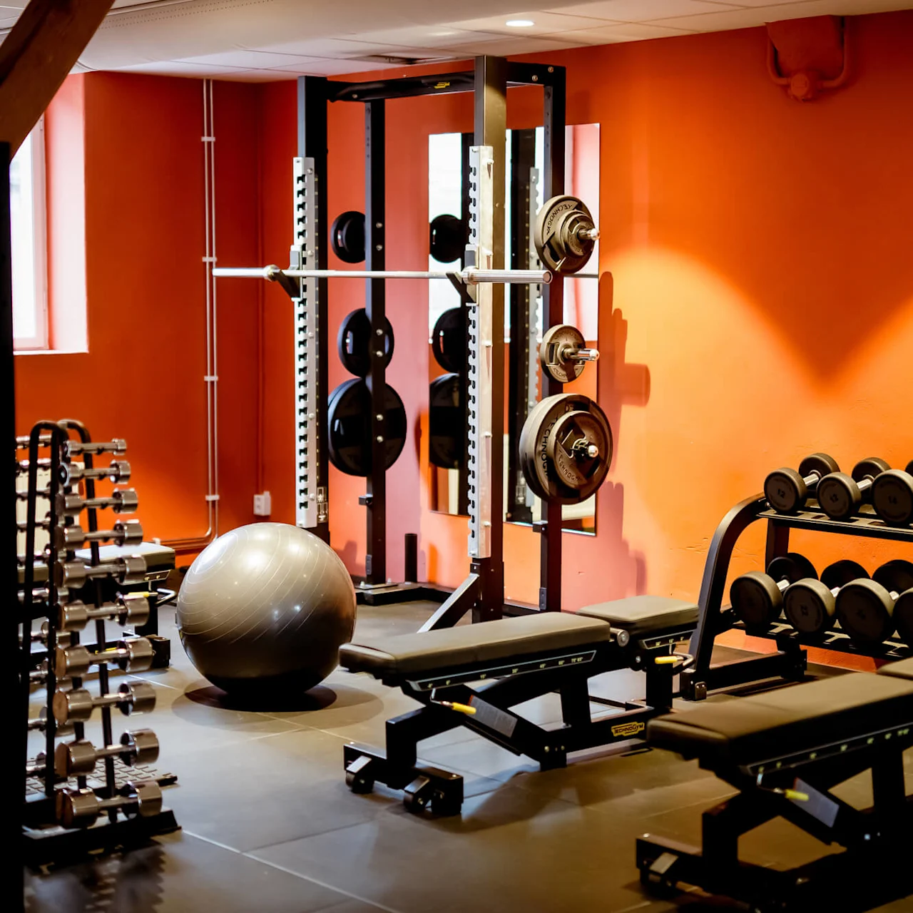 A gym with weights, benches, and a stability ball, signifying fitness activity, in a room with orange walls.