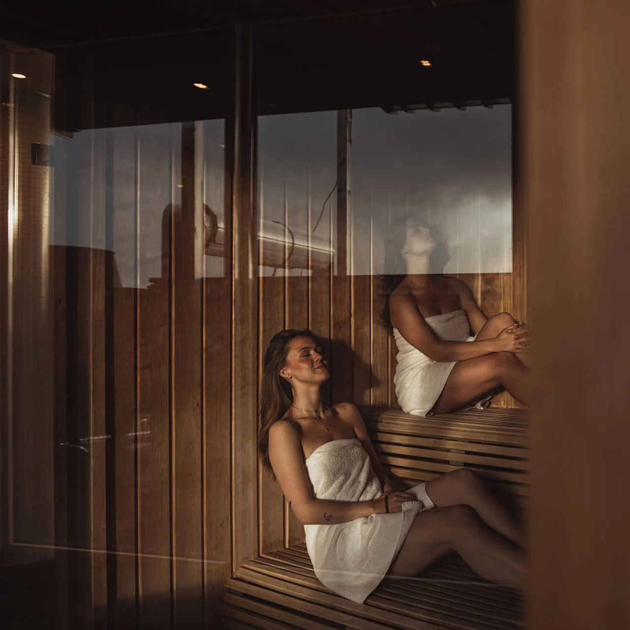 Two women, wrapped in towels, sit relaxing on wooden benches in a dimly lit sauna. Steam rises, creating a tranquil atmosphere.