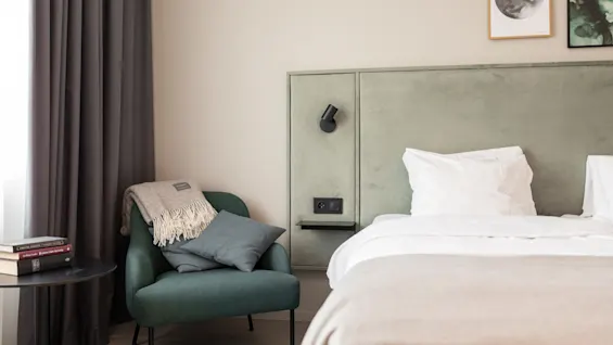 Rauhallinen hotellihuone, jossa on mukava sänky ja viihtyisä lukunurkkaus, täydennettynä nojatuolilla, peitolla ja kirjoilla.