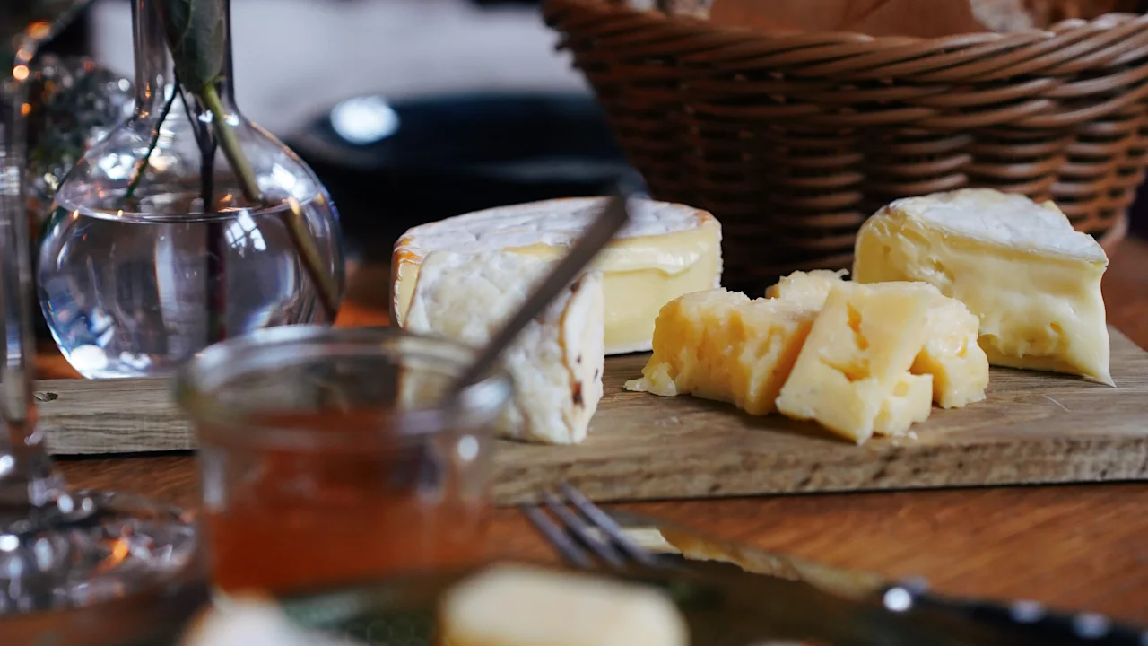 Cheese assortment rests on a wooden board with bread basket and wine nearby; set on a dining table with a vase of foliage in a dimly lit setting.