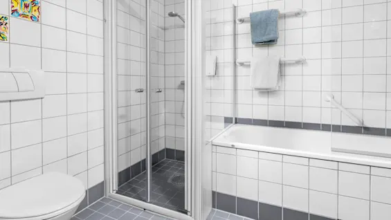 A white-tiled bathroom features a toilet, shower with glass doors, and bathtub. Towels hang on bars above the tub. Vibrant decorative tiles accent the wall.