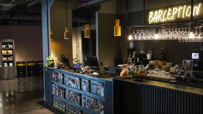 A bar counter doubling as a reception desk with pendant lights, in a dimly lit, modern space with shelves of snacks.