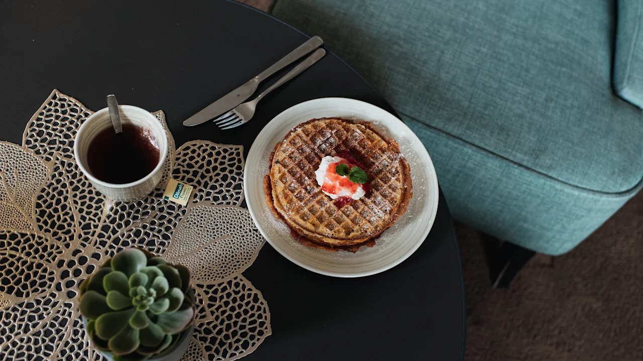 A plate with waffles topped with whipped cream and strawberries on a table. Beside it, a cup of tea with a teabag and a succulent plant sit on a woven placemat.