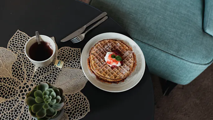En tallerken med vafler toppet med pisket krem og jordbær på et bord. Ved siden av, en kopp te med en tepose og en sukkulent plante på en vevd duk.