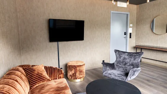 En brun sammetssoffa vetter mot en stor svart TV på en beige vägg, tillsammans med en rund grå stol och ett cirkulärt svart soffbord på trägolv.