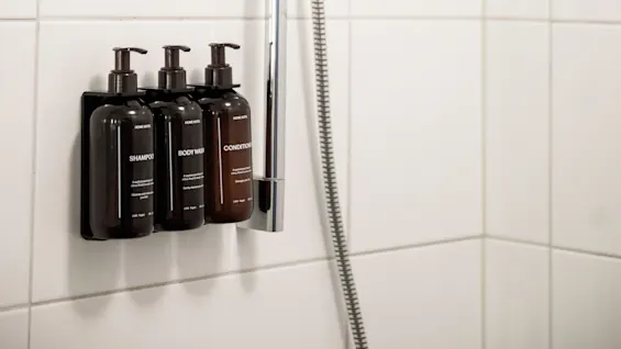Duschhuvud på justerbar kromhållare; tre väggmonterade flaskor märkta SCHAMPOO, BODY WASH, BALSAM; vit kakelvägg i bakgrunden.