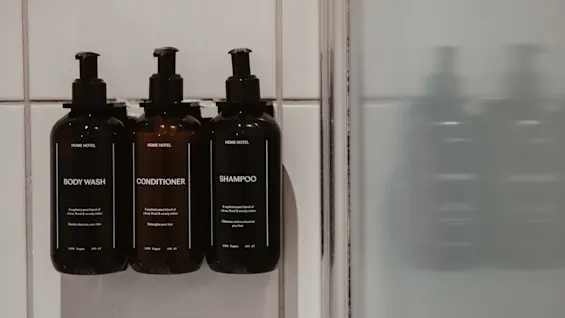 Tre mørke flasker med BODY WASH, CONDITIONER og SHAMPOO på en flisebelagt brusevæg, klar til en forfriskende rens.
