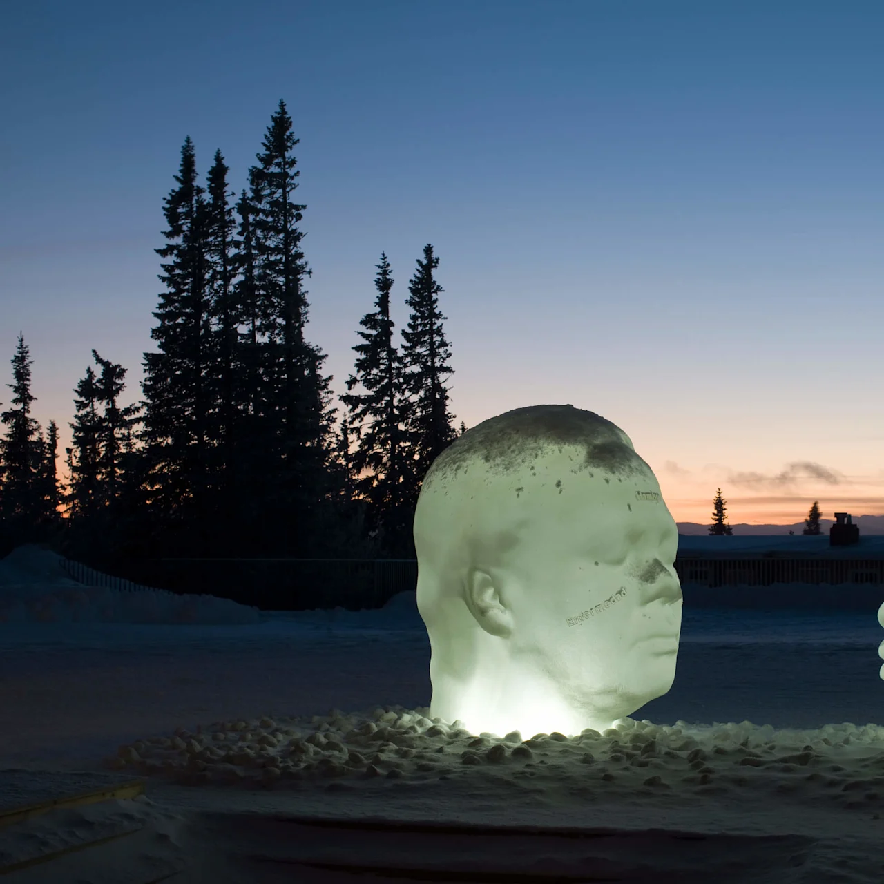 Two illuminated ice sculptures of human heads face each other against a twilight sky with silhouetted trees.