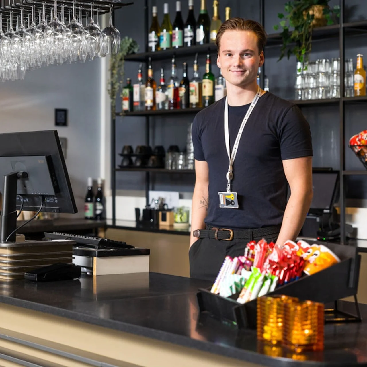 A smiling person stands behind a bar counter. Shelves with bottles and bags of chips fill the background. A basket of snacks and two computer monitors are on the counter.