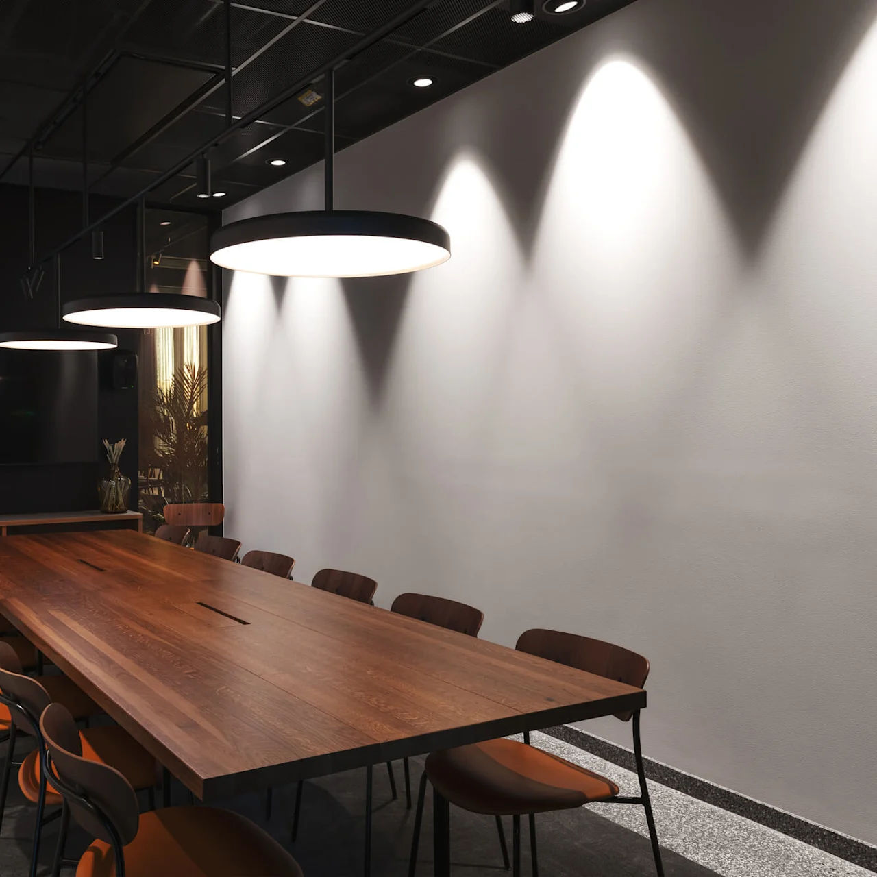 A long wooden conference table with chairs beneath circular ceiling lights; a flat screen and cabinets on a wall suggest an office meeting room setting.