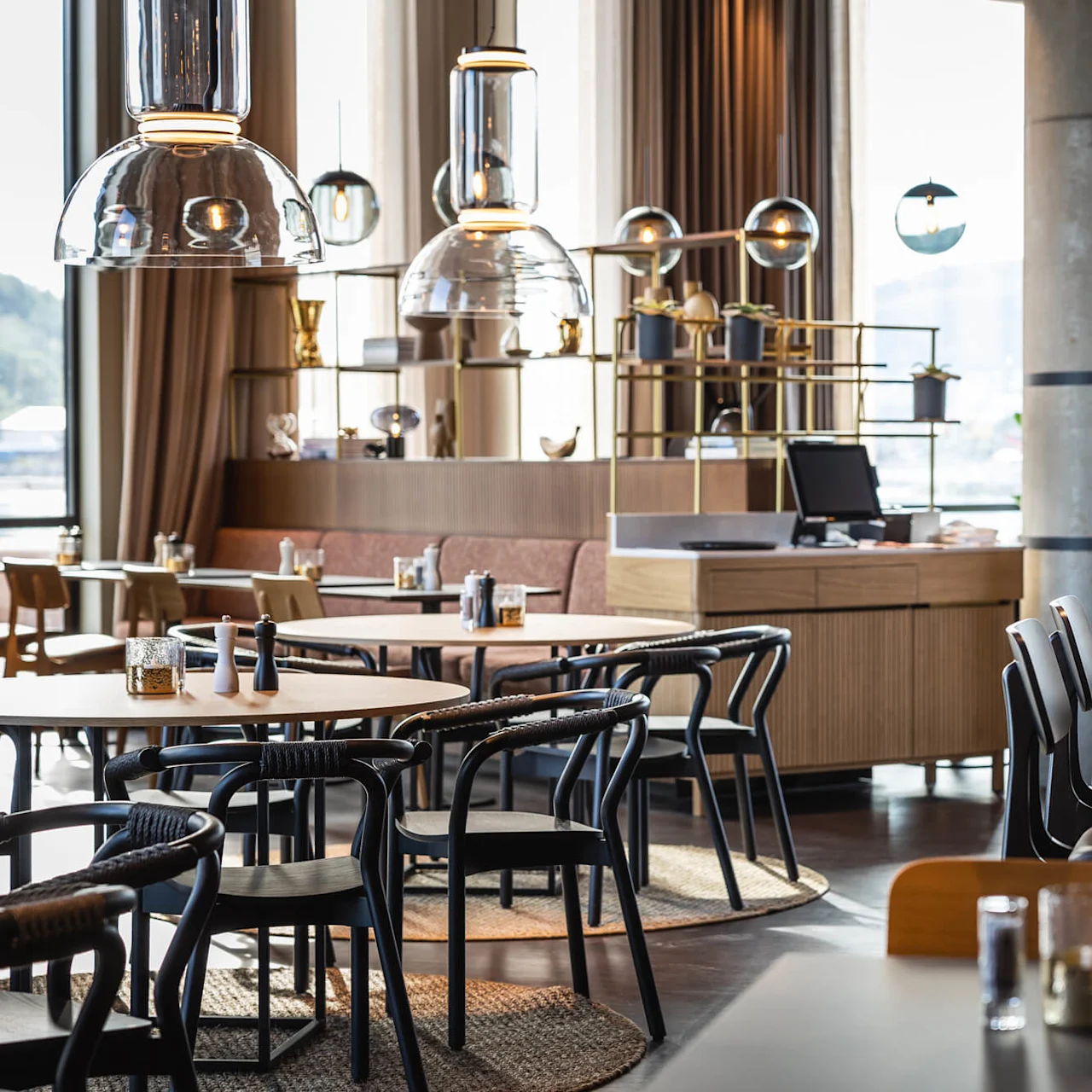 Borde og stole er arrangeret i en moderne restaurant, der har pendellamper og en frodig plantevæg med store vinduer, der viser en bysilhuet i baggrunden.