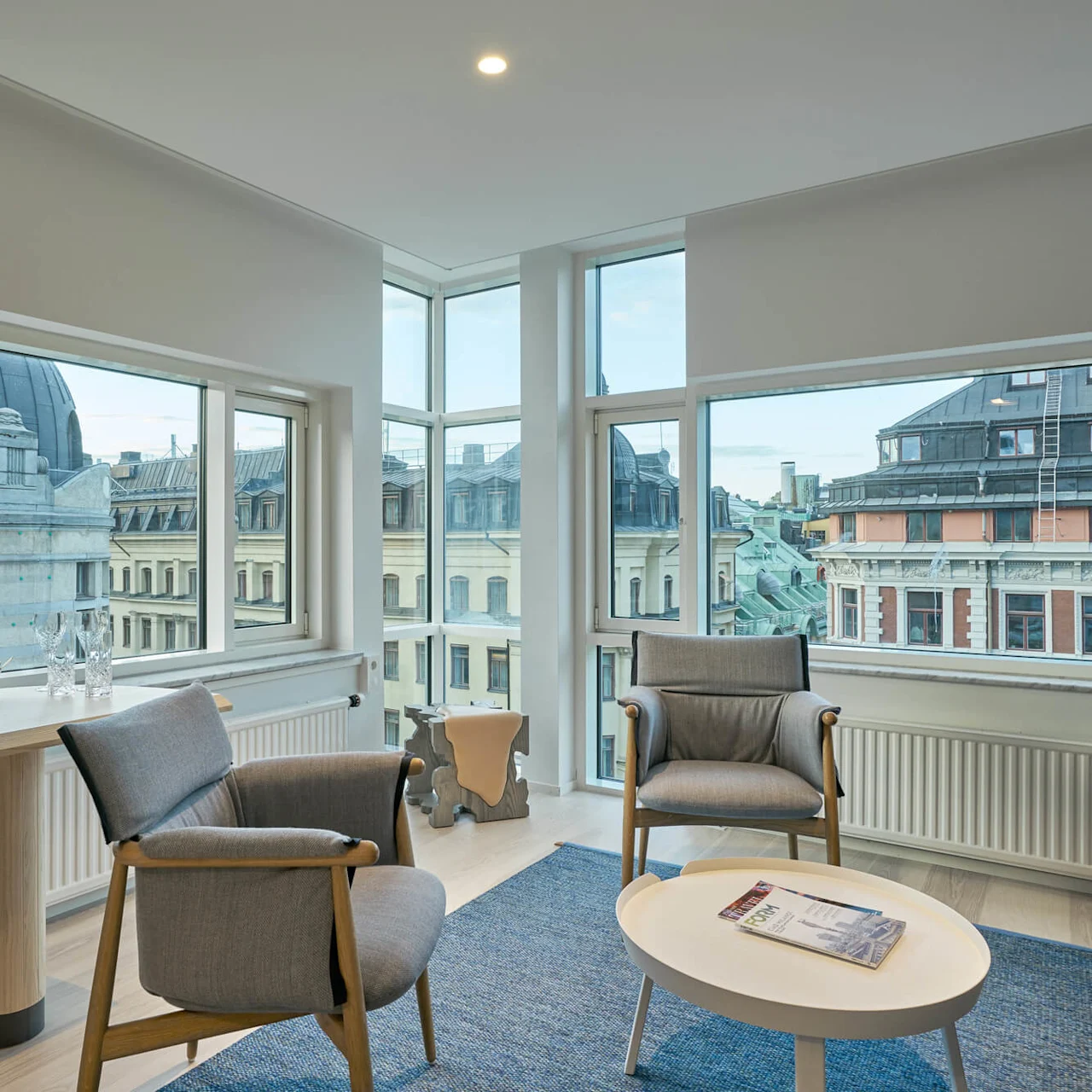 wooden panel wall background chairs sit near a coffee table with a magazine, framed by large windows offering views of adjacent historic buildings under a blue sky.