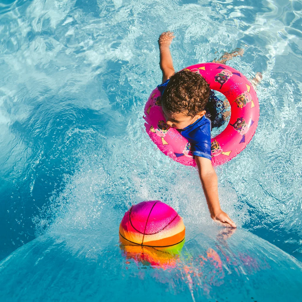 Child in a pink inflatable ring playing with a colorful ball on a water jet in a swimming pool.