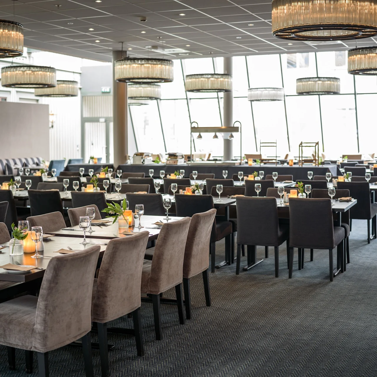 Eleganta matbord är prydligt arrangerade med vinglas och servetter, upplysta av ljuskronor i en rymlig restaurang med stora fönster.
