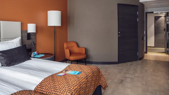 Säng med vita lakan och orangefärgad mönstrad filt vilar bredvid ett runt bord och lampa, i ett modernt hotellrum med en orange stol och trägolv.