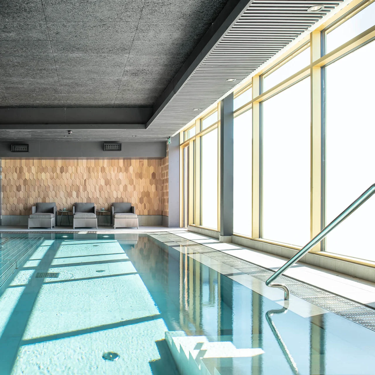 An indoor pool with clear water sits beside three gray lounge chairs. Sunlight filters through large windows on the right, illuminating the space with a relaxing ambiance.