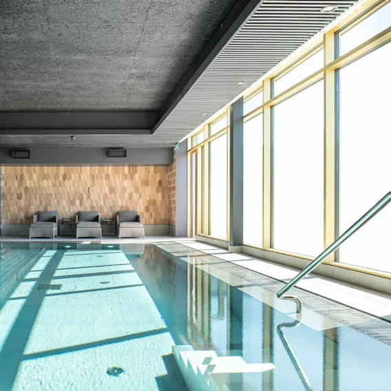 En inomhuspool med klart vatten ligger bredvid tre gråa solstolar. Solljus filtreras genom stora fönster till höger och ger rummet ett avslappnande ljus.
