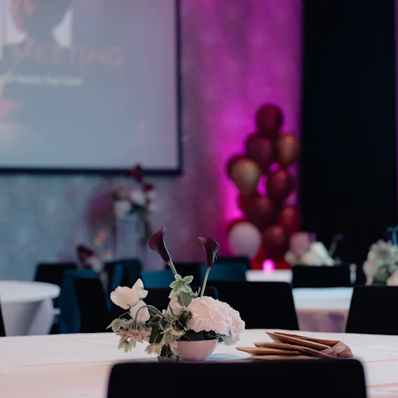 Kokoushuone valmiina CLARION MEETING -tapahtumaan, pöytiä, tuoleja, kukka-asetelmia ja ilmapalloja, valaistu violetilla valolla.