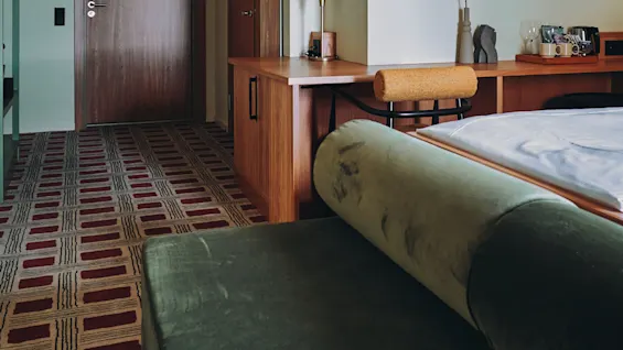 Et koselig hotellrom med en grønn sofa, en seng og et skrivebord. Et mønstret teppe fører mot utgangsdøren.