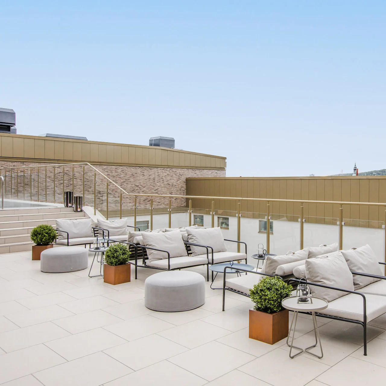 Udendørs siddeområde med metalrammede sofaer med grå puder og små borde, placeret på en flisebelagt tagterrasse med glasrækværk; bjerge og himmel kan ses i baggrunden.