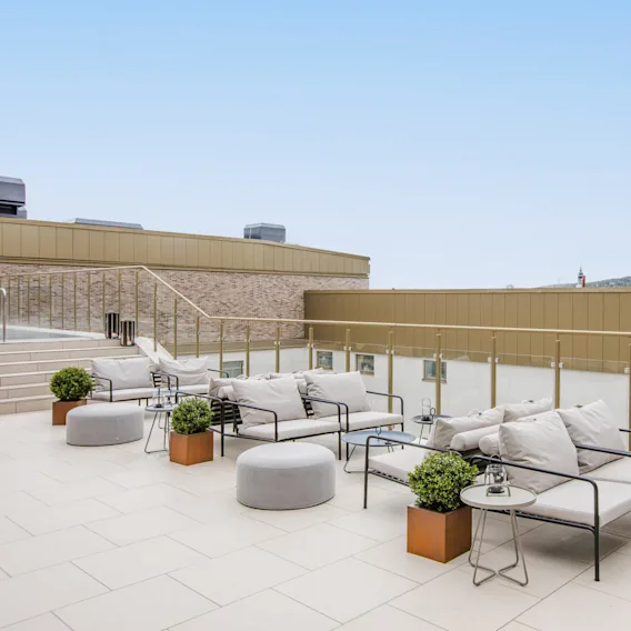 Outdoor seating area featuring metal-framed sofas with gray cushions and small tables, set on a tiled rooftop terrace with glass railings; mountains and sky visible in the background.