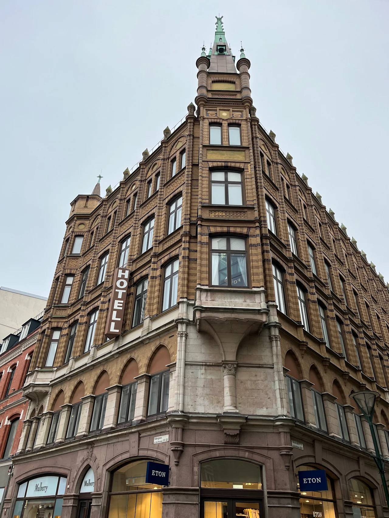 En murbygning med flere etasjer viser ordet "HOTELL" vertikalt på fasaden, og har butikker merket "Mulberry" og "ETON" på bakkeplan.