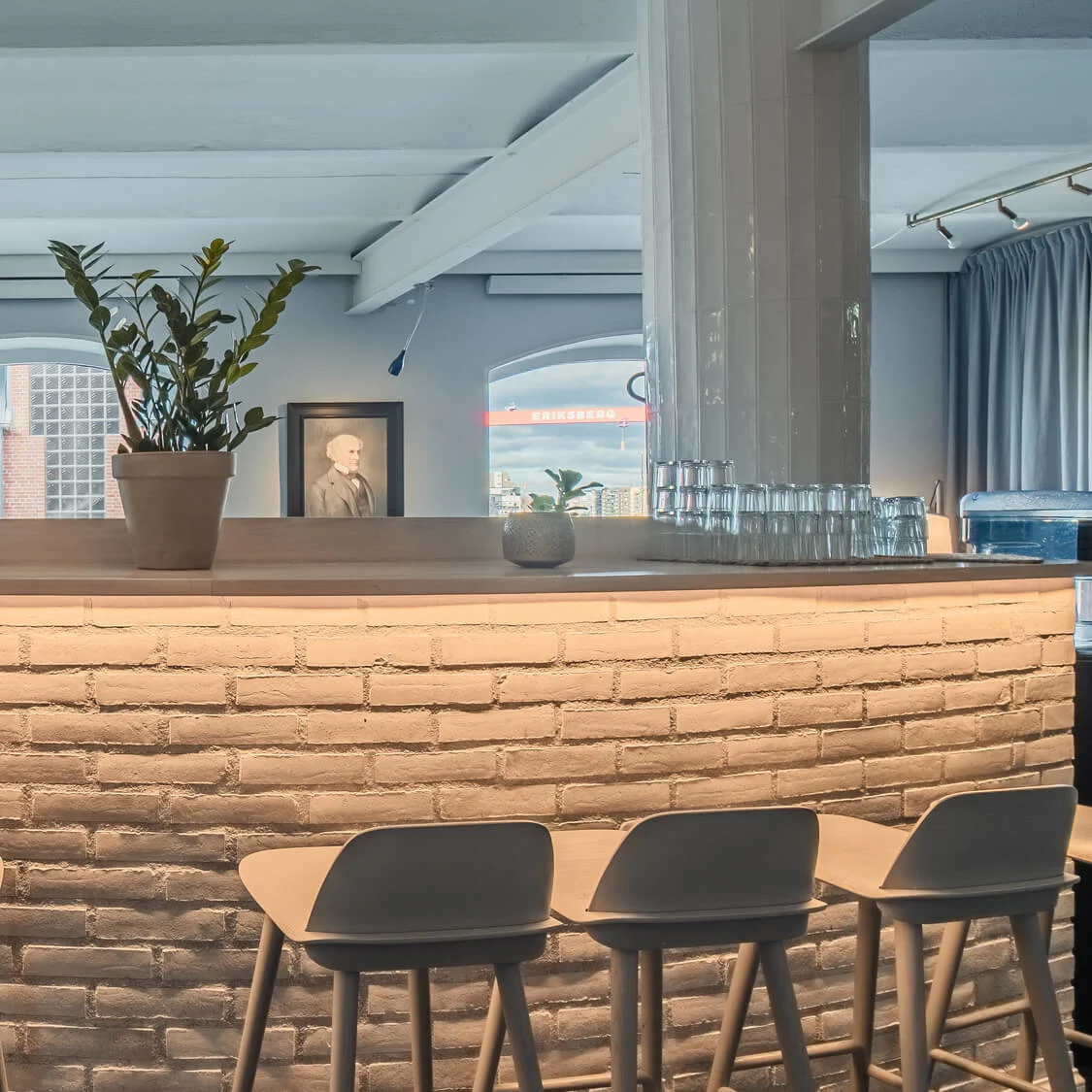 A modern bar counter with six stools stands illuminated in a stylish room. A potted plant and glassware adorn the counter. Windows reveal a cityscape outside. "ERIKSBERG" is visible.