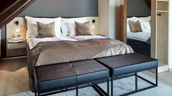 A well-made bed with brown bedding in a cozy, wood-accented bedroom with sloped ceilings and a reflective chandelier.