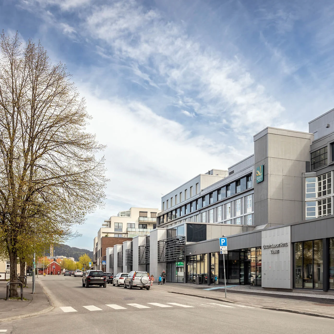 En moderne, fler-etagers hotelbygning står ved en gade med parkerede biler og træer. Fire flagstænger står foran. Teksten lyder: "Quality Airport Hotel Værnes."