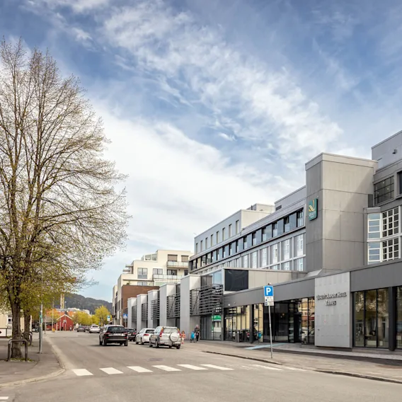 En moderne fleretasjes hotellbygning ligger ved en gate med parkerte biler og trær. Fire flaggstenger står foran. Teksten lyder: "Quality Airport Hotel Værnes."