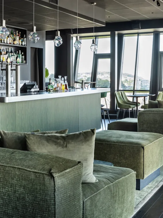 En moderne bar har grønne, polstrede sitteplasser, en elegant disk med hengende lys og hyller fylt med flasker. Store vinduer gir utsikt til et fjernbybilde.
