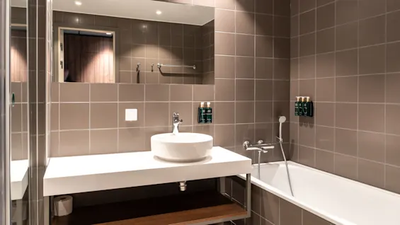 Ett modernt badrum med ett vitt handfat på en svävande bänk bredvid ett badkar, grå kaklade väggar och en stor spegel som reflekterar det rena, minimalistiska designen.