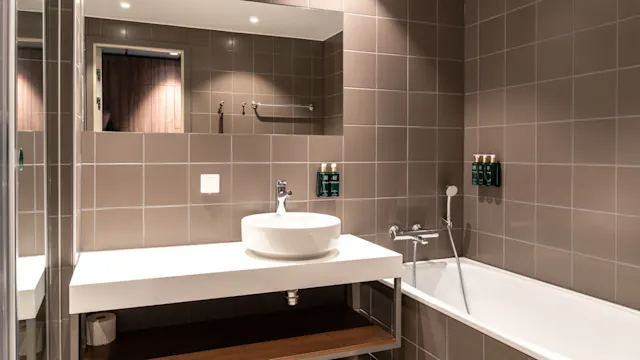 Et moderne badeværelse med en hvid vask på en svævende bordplade ved siden af et badekar, grå flisevægge og et stort spejl, der reflekterer rummets rene, minimalistiske design.