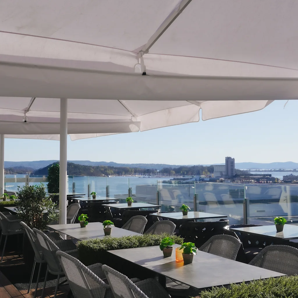 A sunny outdoor terrace with tables and chairs under large umbrellas, overlooking a scenic bay and city.