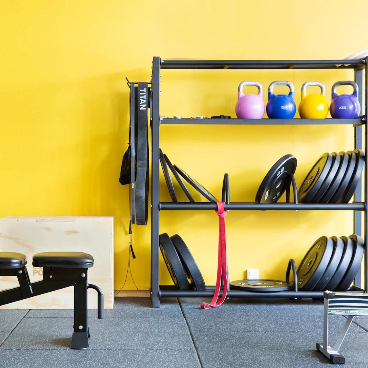 En træningsbænk står nær en reol med kettlebells og vægtskiver mod en lys gul væg. En trækasse og modstandsbånd giver flere træningsmuligheder.