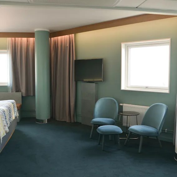 A bedroom features a bed and two chairs with a small table in a corner. Windows allow natural light, while curtains and a television complete the modern, minimalistic setting.