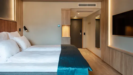 Ett rymligt hotellrum med en prydligt bäddad dubbelsäng med ett teal täcke och väggbelysning, trägolv, platt-tv och en dörr som leder ut.