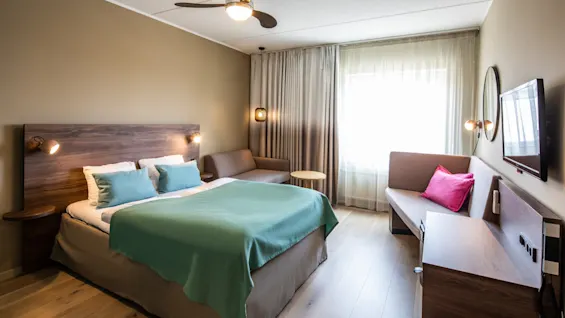 Bed positioned centrally with teal bedding; wall-mounted lamps illuminate both sides. Nearby, a couch accessorized with a pink cushion sits under sheer-draped window. Wooden floor and ceiling fan complete the cozy room.