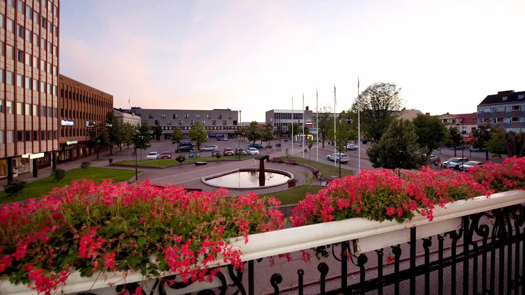 En stadsvy i skymningen med blommande växter i förgrunden, en fontän i mitten av ett torg, omgiven av byggnader, gator och parkerade bilar.